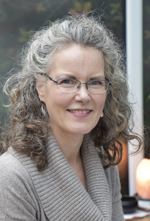 An image of me; a woman in her 50s with long, brown greying curls and rimless glasses. I am looking into the camera and smiling.